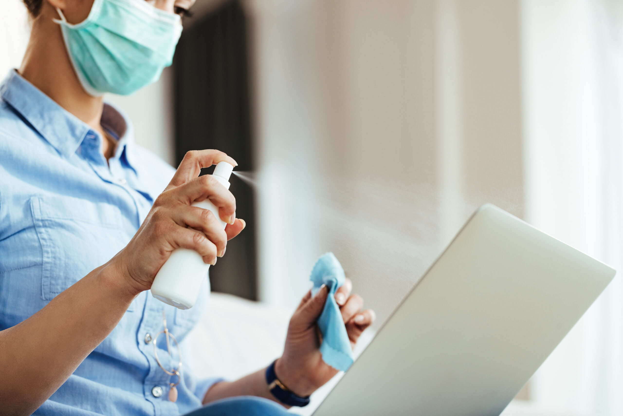 Close-up of woman using spray bottle while cleaning her laptop during virus epidemic.